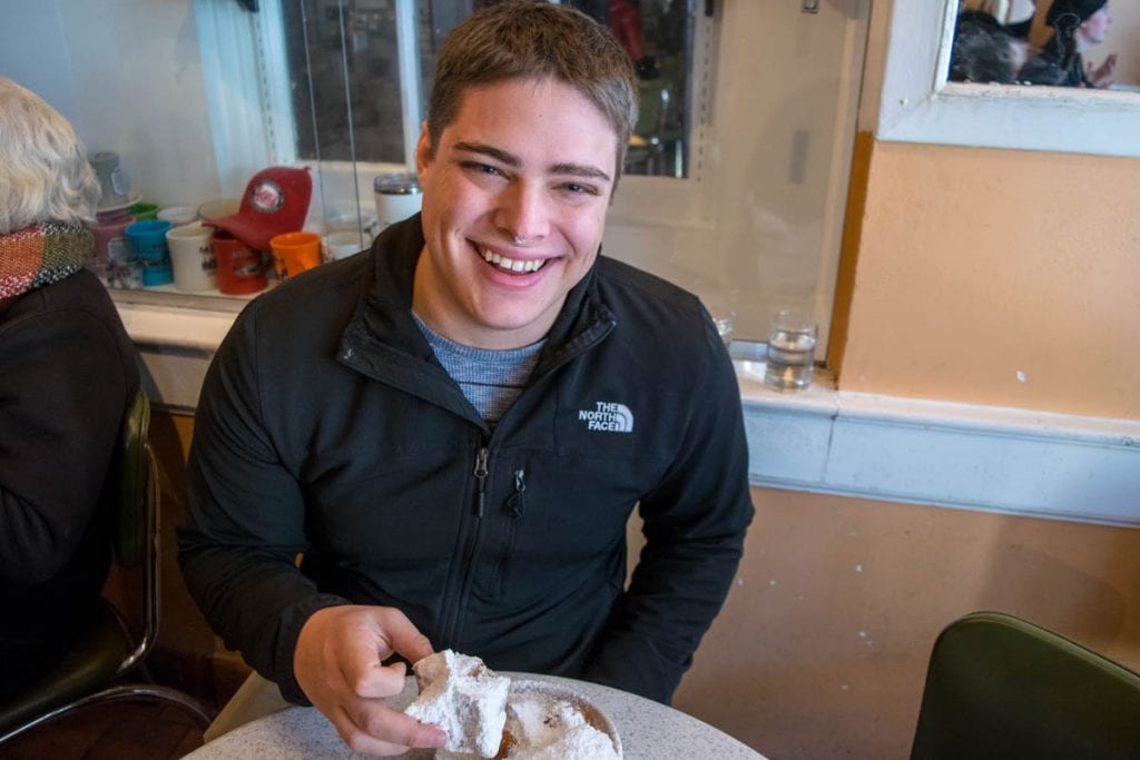Jeremy Storm in a black jacket holding a beignet in Cafe du Monde during a trip to New Orleans Louisiana