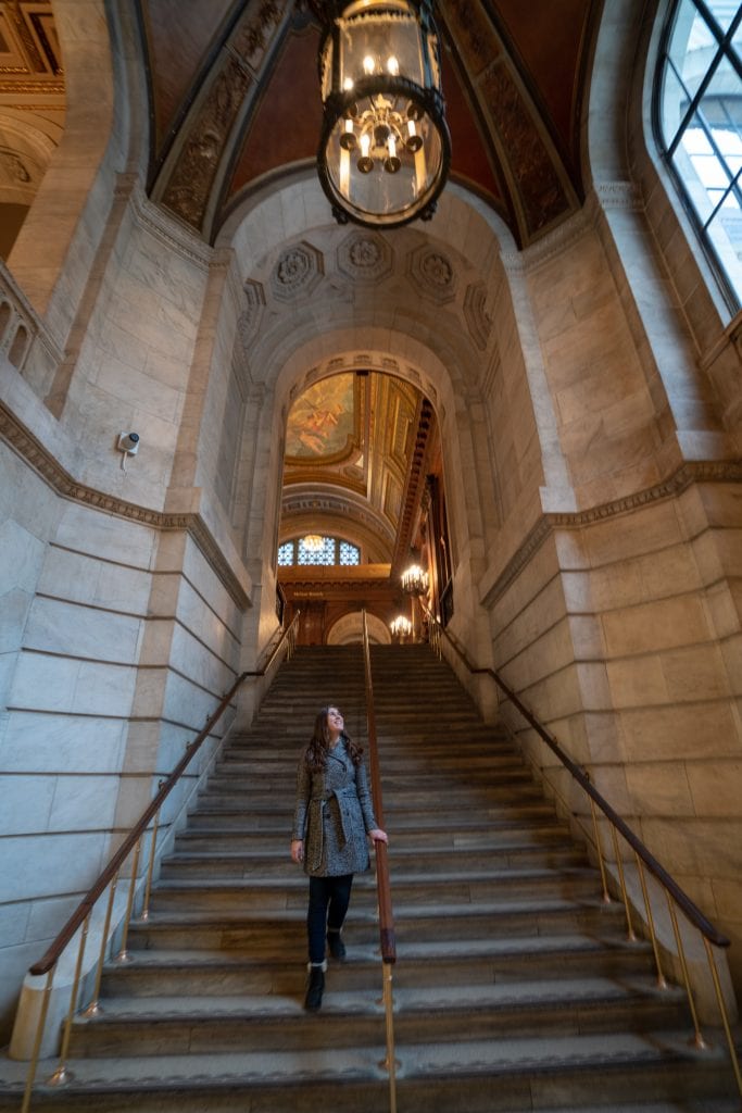 Kate storm walking down a stone staircase in the NYC Public Library, 5th Avenue branch. Kate is wearing a gray coat.