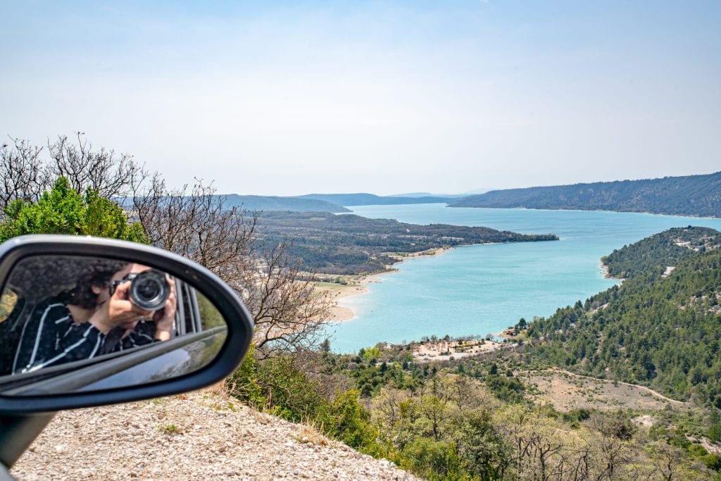 Photo of Lake Sainte-Croix with Kate visible snapping a photo in the rearview mirror on the left--a camera for photos like this definitely belongs on your road trip packing list!