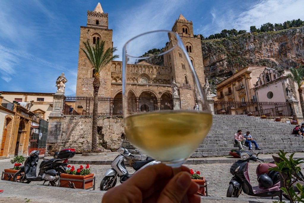 glass of white wine being held in front of church in cefalu sicily