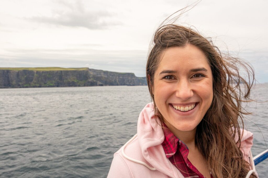 Kate Storm wearing a pink jacket and smiling at the camera during a Cliffs of Moher cruise--the cliffs as visible in the background.