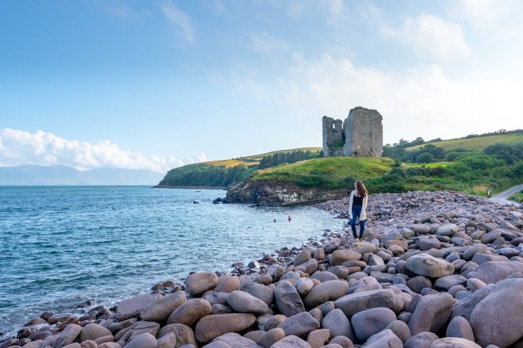 Kate Storm standing on a pebble beach on Dingle Peninsula, Ireland. Minard Castle is behind her. This is a great example of what to wear in Ireland!