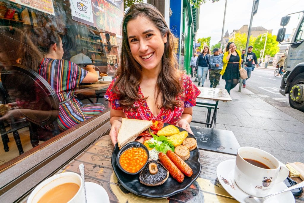 Kate Storm in a red dress holding up a full Irish breakfast while sitting at a small table on the sidewalk in Dublin