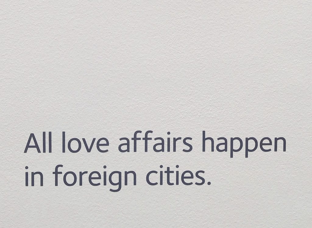 Photo of white wall in the Museum of Broken Relationships in Zagreb Croatia. Black text reads "All love affairs happen in foreign cities."
