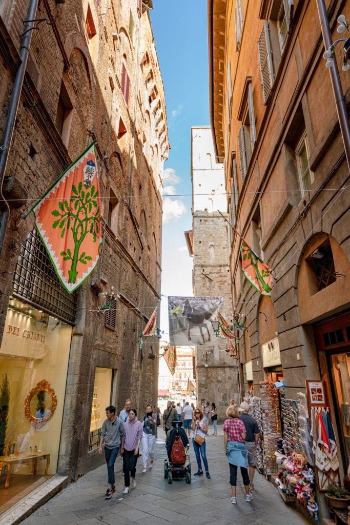 narrow street in siena italy with contrade flag hanging from the buildings on the left, a fun stop during a 3 day tuscany itinerary