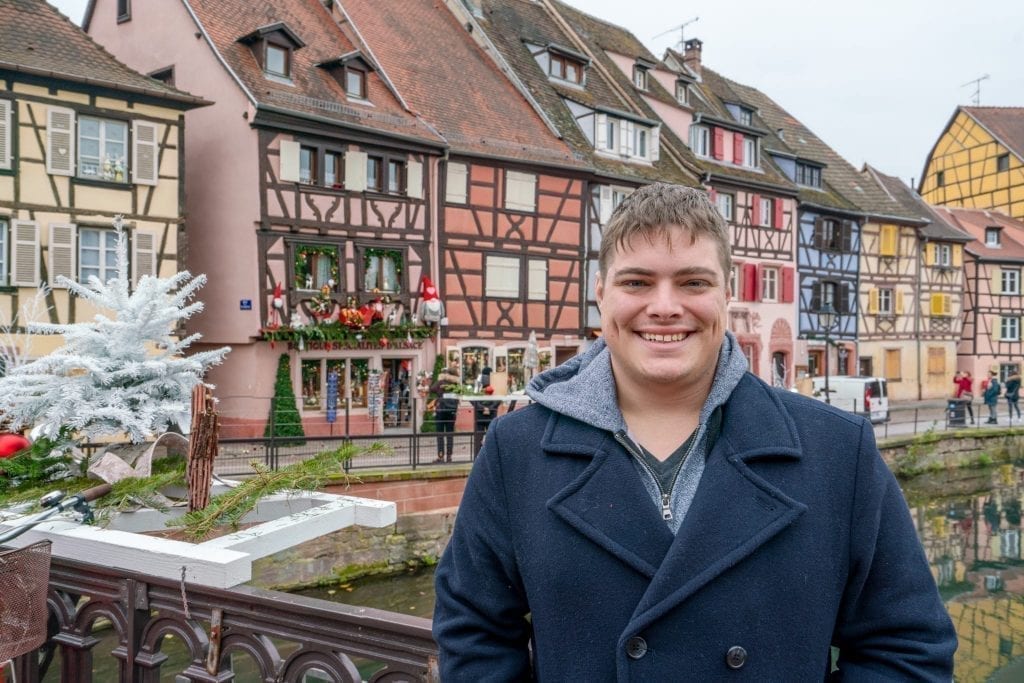 Jeremy Storm in a blue coat standing in front of colorful houses in Colmar France