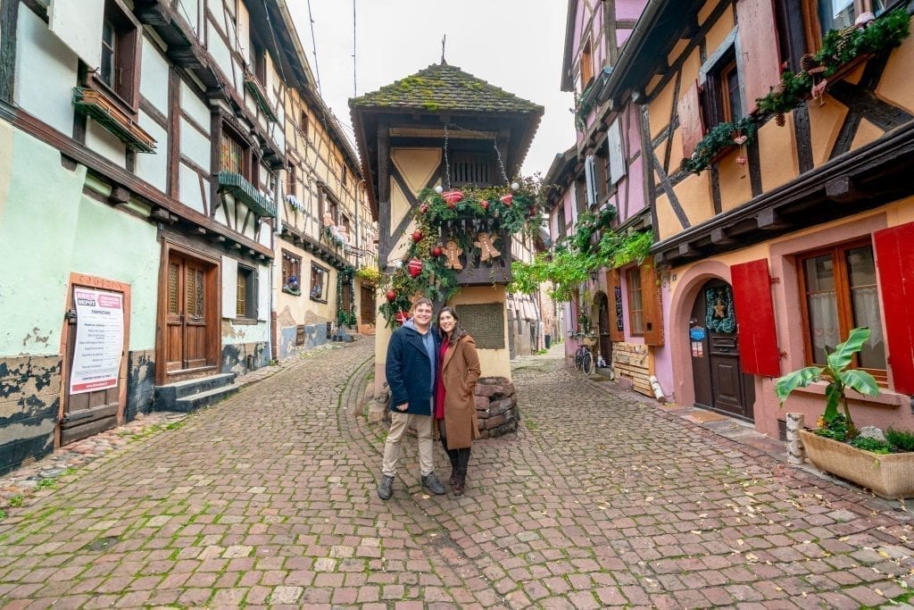 Kate Storm and Jeremy Storm wearing coats and standing in Eguisheim France surrounded by half-timbered houses