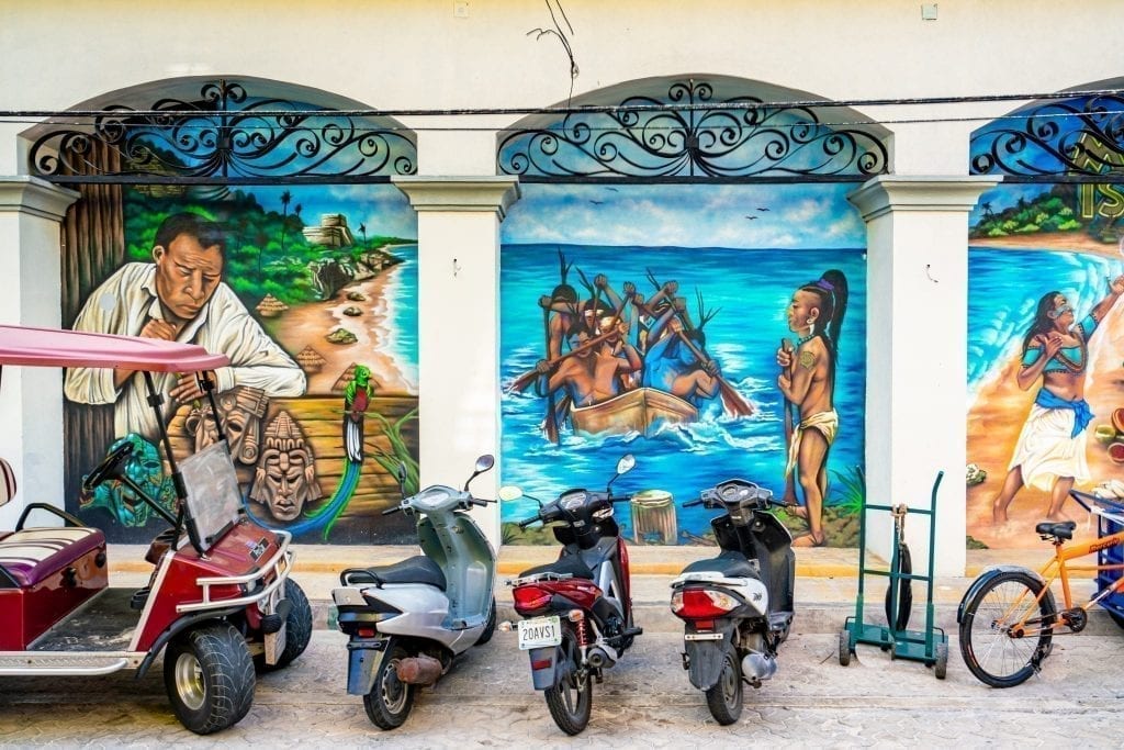 Street art in Isla Mujeres Mexico with mopeds and a golf cart parked in front of the building it is on. The street art depicts Mayan culture.