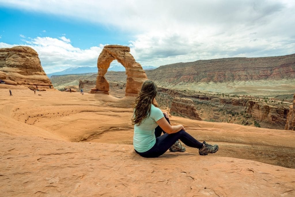 Kate Storm sitting in front of Delicate Arch Utah wearing a turquoise shirt