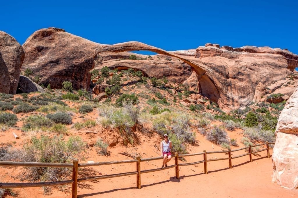 Kate Storm sitting on a fence in front of Landscape Arch, one of the best hikes in Arches National Park