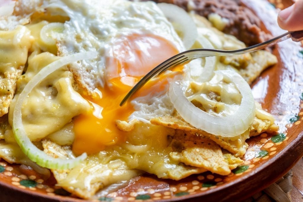 Pile of chilaquiles with green sauce topped with a fried eggs with a fork about to dive in. Chilaquiles are a typical Mexican breakfast dish.