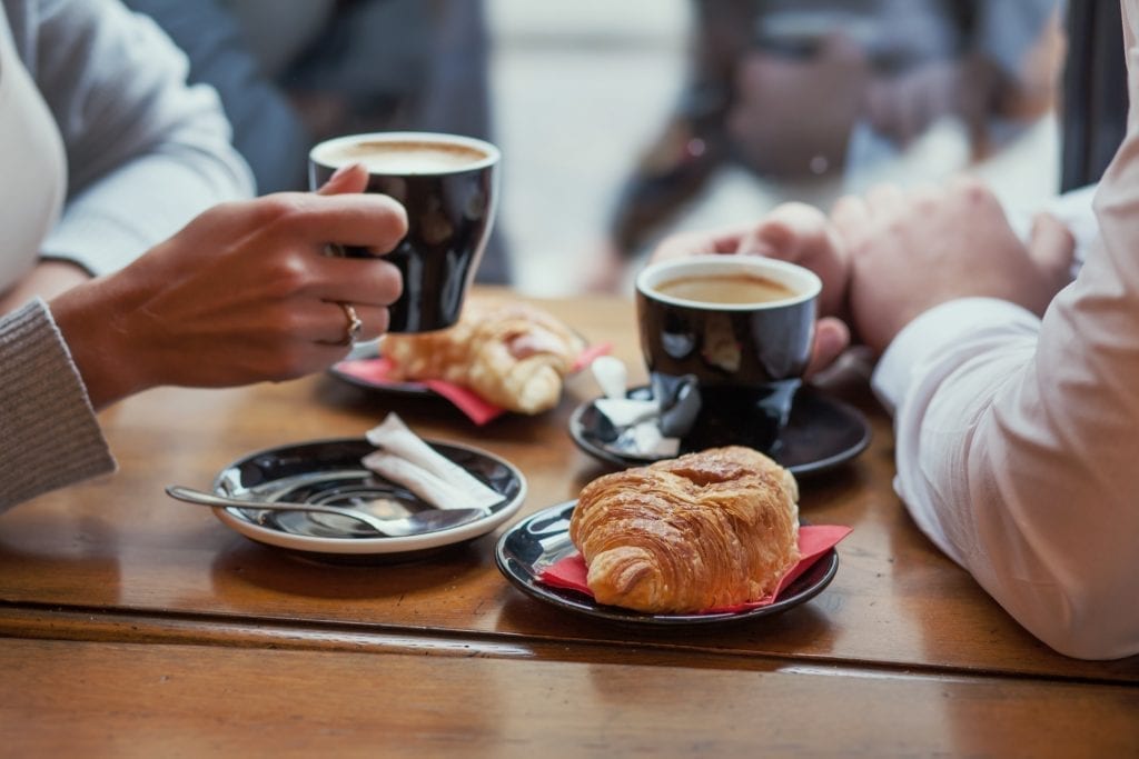 2 people holding coffee cups over a brown table in France with croissants by their sides. The focus of the image is the food.