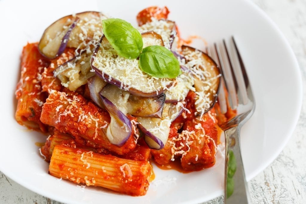 Pasta alla Norma topped with eggplant in a white bowl with a silver fork next to it