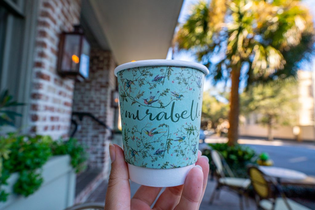 colorful blue cup that says "mirabelle" being held outside of mirabelle cafe, one of the best savannah coffee shops