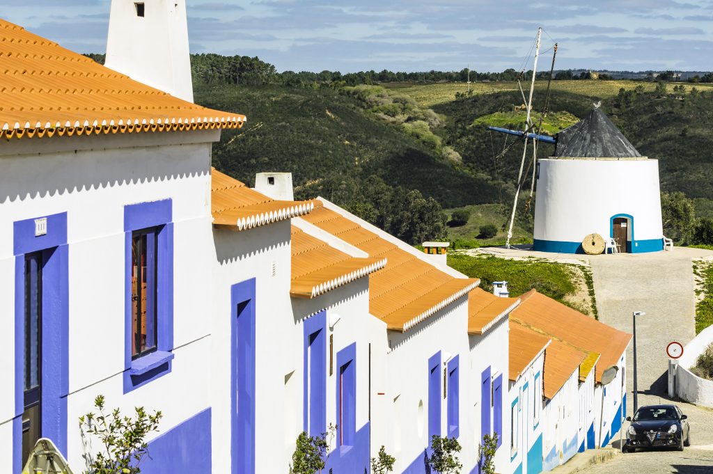 white buildings with purple trim and a windmill in the background in odeceixe portugal