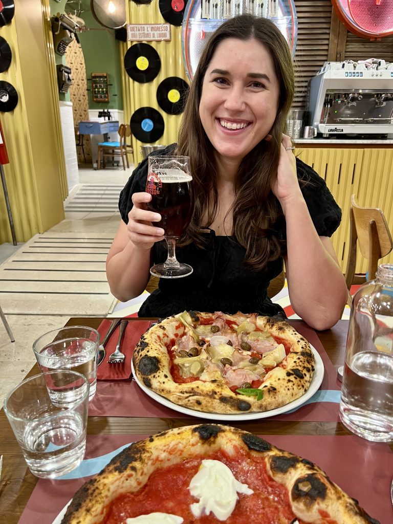 kate storm with a capricciosa traditional pizza in italy holding up a beer
