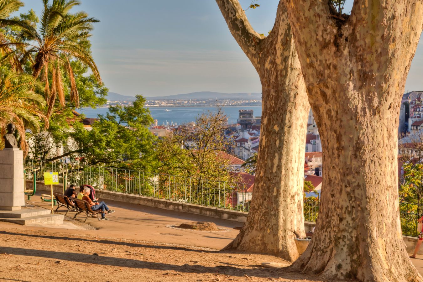 miradouro do torel pictured with trees in the foreground and people looking out over one of the best views of lisbon portugal from a bench