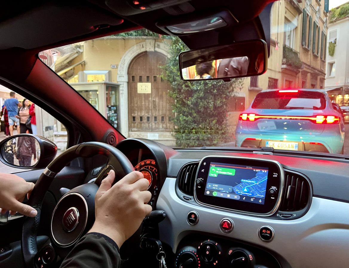 view of front dash of car and jeremy storm's hands driving through sirmione historic center