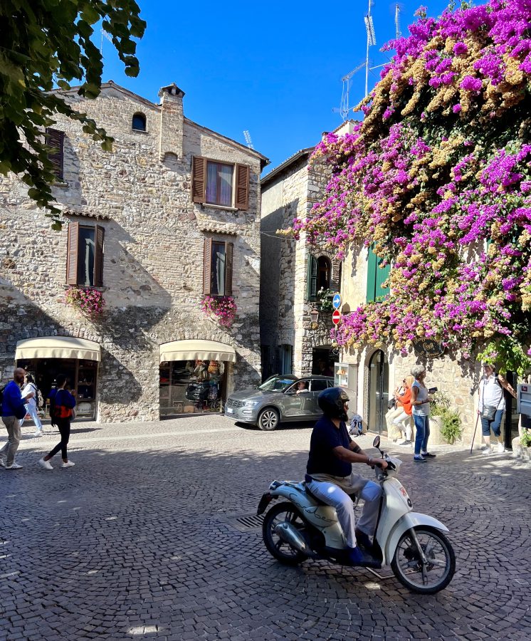 person on a motorbike crossing through a piazza in sirmione with a bougainvillea blooming in the background