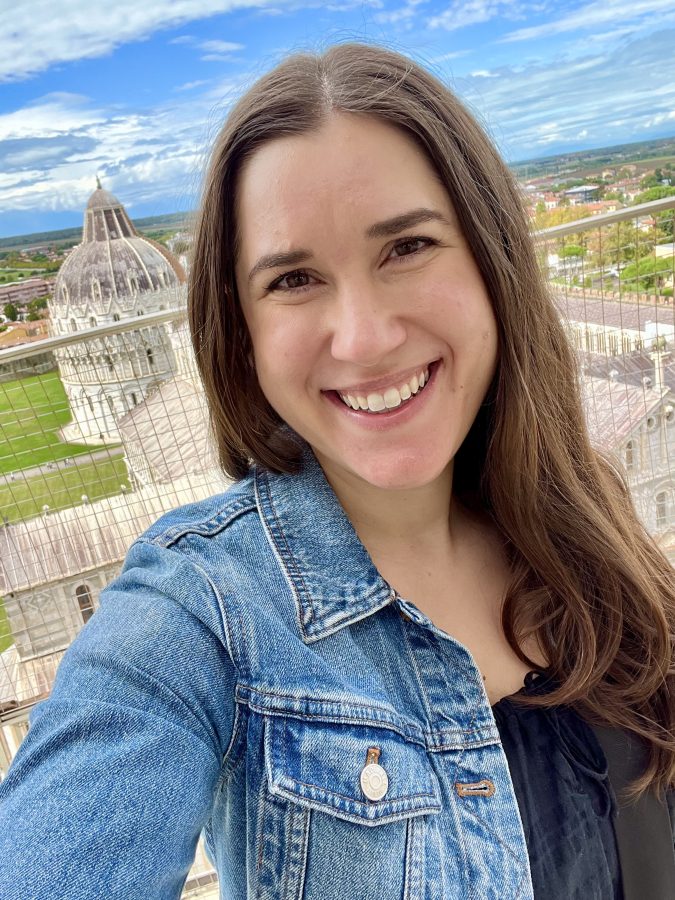 kate storm taking a selfi at the top of the leaning tower of pisa with fence and part of the baptistery visible behind her
